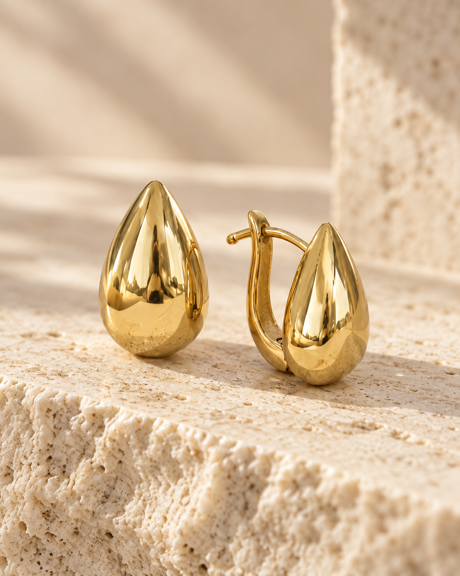 Pair of teardrop huggie earrings on a stone ledge, mirror-finish gold teardrops with hinged latch wires